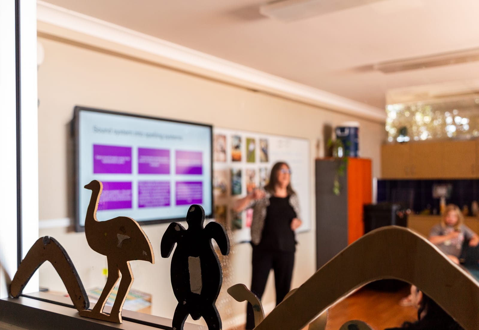 A facilitator presenting to community members during a Gathang language session, with carved emu, turtle and wave figures in the foreground